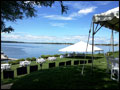 white large tents and tables set for a catered wedding, on a green lawn overlooking water